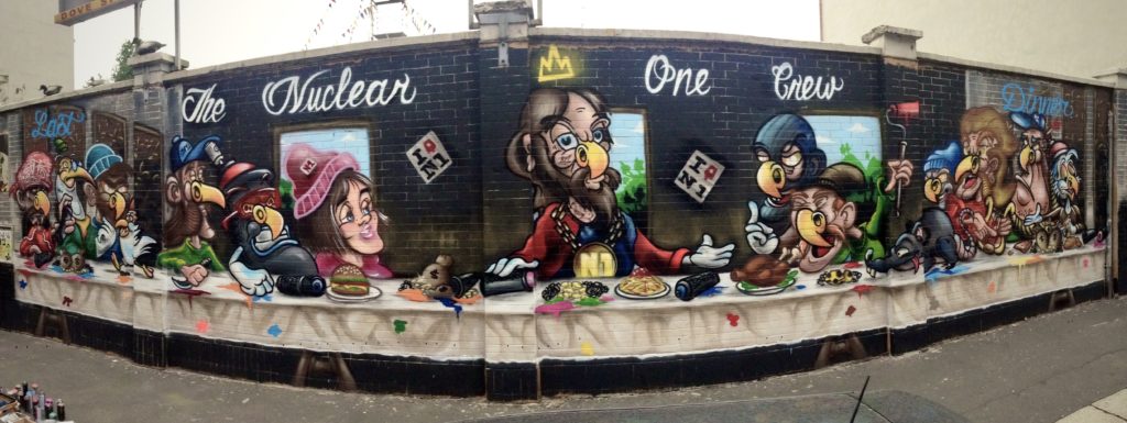 Murales in stile graffiti con figure caricaturali su un muro esterno realizzato dal collettivo Nuclear Crew